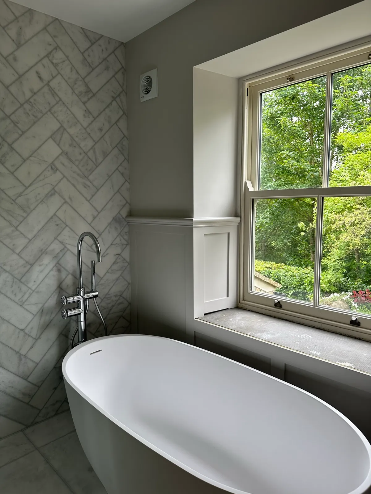 A freestanding bath beside a tiled wall and sash window.